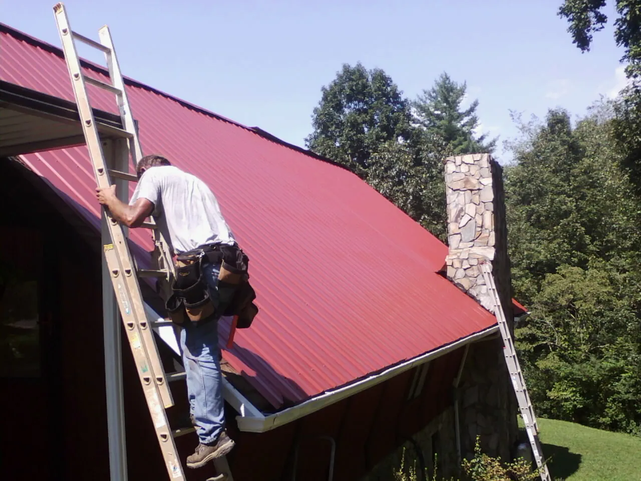 Roofer on a ladder beside a red metal roof and stone chimney — Soffit & Fascia Repair by Roofing Brothers in Shipman